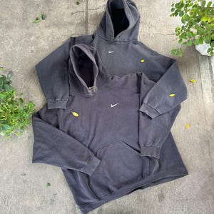 May include: Two dark gray hoodies with the Nike logo, one on top of the other. The hoodies have a front pocket and a drawstring hood. The hoodies are on a concrete surface with some leaves and greenery.
