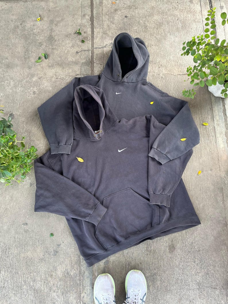 May include: Two dark gray hoodies with the Nike logo, one on top of the other. The hoodies have a front pocket and a drawstring hood. The hoodies are on a concrete surface with some leaves and greenery.