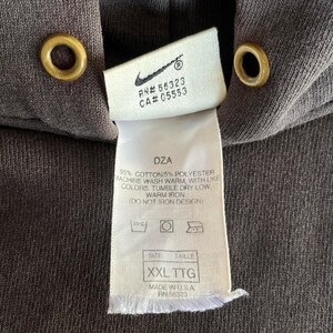 May include: Close-up of a dark brown hoodie with a tag. The tag features the Nike logo, RN and CA numbers, and washing instructions. The size is XXL TTG. The hoodie has gold-colored eyelets.