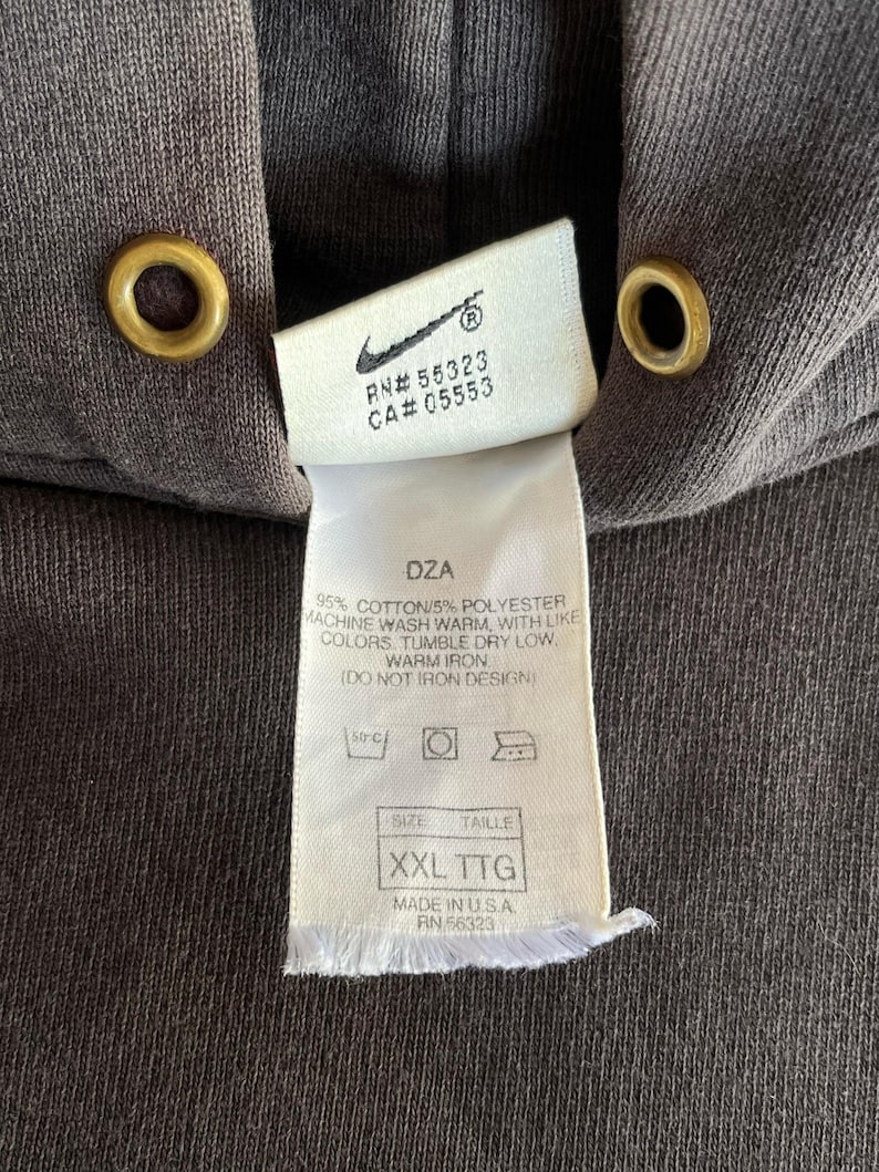 May include: Close-up of a dark brown hoodie with a tag. The tag features the Nike logo, RN and CA numbers, and washing instructions. The size is XXL TTG. The hoodie has gold-colored eyelets.