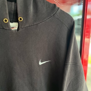 May include: Dark gray Nike hoodie with a white Nike swoosh logo on the chest. The hoodie has a drawstring hood with gold-colored eyelets. The garment is hanging on a wooden hanger.