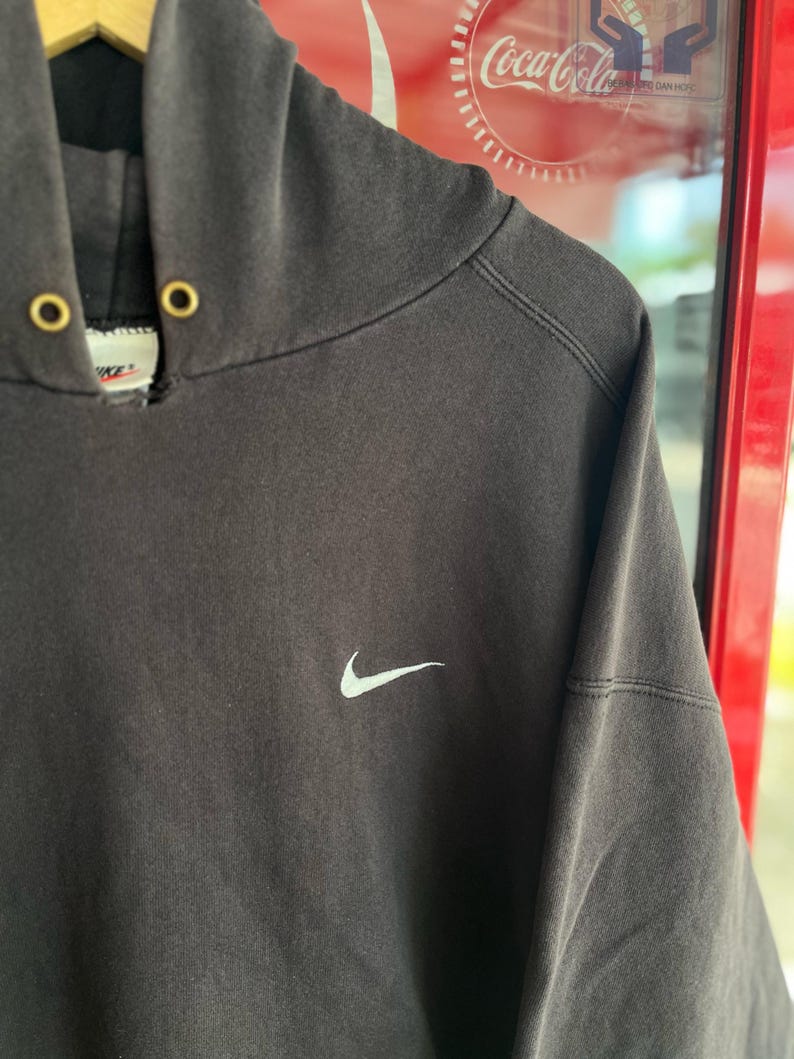 May include: Dark gray Nike hoodie with a white Nike swoosh logo on the chest. The hoodie has a drawstring hood with gold-colored eyelets. The garment is hanging on a wooden hanger.