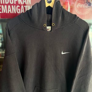 May include: A dark gray Nike hoodie with a white Nike swoosh logo on the chest. The hoodie has a drawstring hood, a kangaroo pocket, and gold-colored eyelets. The hoodie is hanging on a wooden hanger.