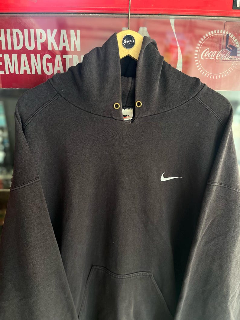 May include: A dark gray Nike hoodie with a white Nike swoosh logo on the chest. The hoodie has a drawstring hood, a kangaroo pocket, and gold-colored eyelets. The hoodie is hanging on a wooden hanger.
