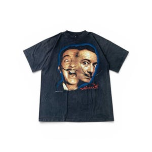 Könnte beinhalten: Dunkelgraues T-Shirt mit einem surrealistischen Porträt von Salvador Dalí. Das Kunstwerk zeigt zwei Gesichter mit einem gemeinsamen Schnurrbart, vor einem blauen Umriss. Die Signatur des Künstlers ist rot.