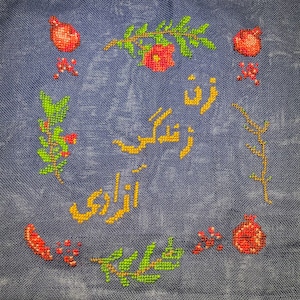 Puede incluir: Textil bordado a punto de cruz con fondo azul. El diseño presenta granadas rojas, elementos florales verdes y rojos, y escritura persa amarilla. El texto está centrado dentro de un borde de motivos florales y frutales.
