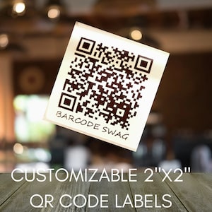 May include: A white square label with a black QR code and the text "BARCODE SWAG". The label is floating in front of a blurred background. The text below the label reads "CUSTOMIZABLE 2"X2" QR CODE LABELS".