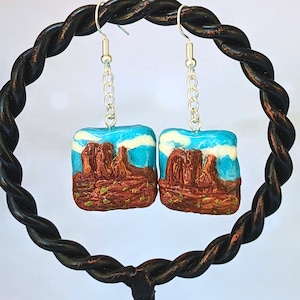 May include: A pair of earrings featuring a blue sky with white clouds and a red rock formation. The earrings are made of polymer clay and have silver hooks.