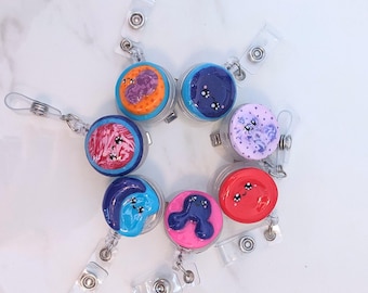 May include: Seven colorful, round, plastic badge holders with cartoon faces and clear retractable badge reels. The badge holders are decorated with various patterns and colors, including red, blue, pink, purple, orange, and white.