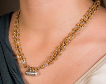 May include: A layered necklace featuring small, round, light brown beads strung on silver-colored chains. A silver-colored cylindrical pendant hangs from the center. The necklace is worn around the neck.