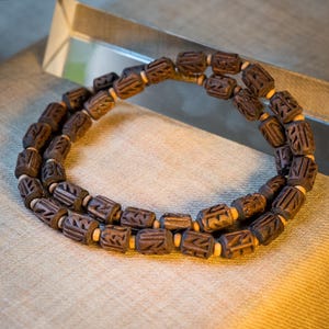 Puede incluir: Un collar de cuentas de madera marrón con intrincados tallados en cada cuenta. El collar está hecho de dos hileras de cuentas y está sobre una superficie de tela beige.