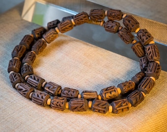 May include: A brown wooden beaded necklace with intricate carvings on each bead. The necklace is made of two strands of beads and is lying on a beige fabric surface.