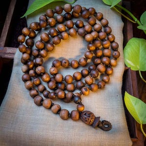 May include: A brown wooden mala bead necklace with a carved wooden pendant. The necklace is made of 108 beads and is strung on a brown cord.