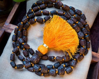 May include: A dark brown wooden mala necklace with a carved design on each bead. The necklace has a bright orange tassel.