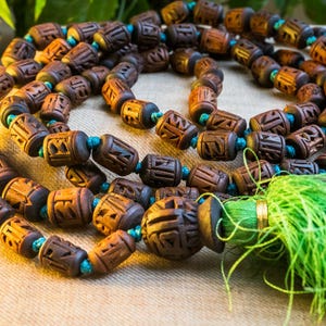 Puede incluir: Un collar de cuentas de mala de madera marrón con un borla verde. Las cuentas están talladas con diseños intrincados y están enhebradas en un cordón azul.