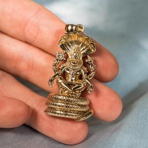 May include: A gold-toned pendant featuring a multi-armed deity figure seated in a cross-legged position atop a coiled serpent base. The figure is framed by a multi-headed cobra hood and holds circular emblems in its upper hands.