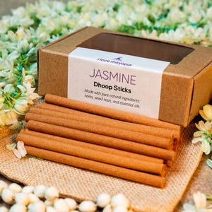 Puede incluir: Una caja de inciensos marrones etiquetados "I love mayapur" y "JASMINE Dhoop Sticks". La caja es de cartón y tiene una ventana transparente en la parte delantera. Los palitos están hechos con ingredientes naturales, incluyendo hierbas, resina de madera y aceites esenciales.