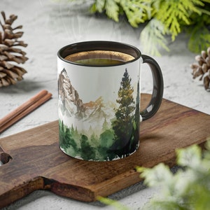 May include: A white ceramic mug with a black interior and handle, featuring a watercolor-style illustration of a mountain range and a dense evergreen forest. The mug is filled with a dark liquid and is positioned on a wooden cutting board.