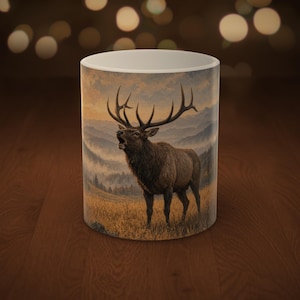 May include: A white ceramic mug featuring a detailed illustration of a large elk with prominent antlers standing in a grassy field. The background depicts a misty mountain landscape during a sunset, with soft golden and blue hues.