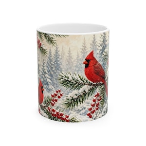May include: A white ceramic mug featuring a winter scene with two bright red cardinal birds perched on snow-covered pine branches. The design includes clusters of red berries and a background of frosted evergreen trees in a snowy forest.