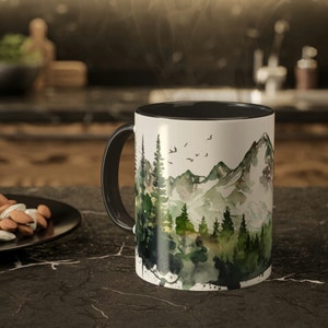 May include: A white ceramic mug with a black interior and handle, featuring a watercolor-style illustration of a mountain range surrounded by a dense forest of green pine trees and small birds flying in the sky above the peaks.