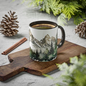 May include: A white ceramic mug with a black interior and handle, featuring a watercolor-style illustration of a mountain range with snow-capped peaks and a dense forest of green pine trees at the base, filled with a dark brown liquid.