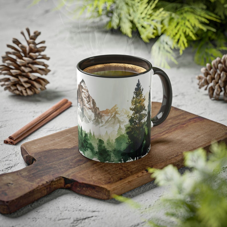 May include: A white ceramic mug with a black interior and handle, featuring a watercolor-style illustration of a mountain range and a dense evergreen forest. The mug is filled with a dark liquid and is positioned on a wooden cutting board.