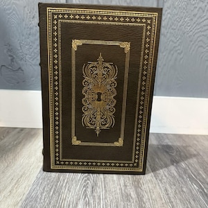 Peut inclure: Un livre marron relié en cuir avec des détails en filigrane dorés. La couverture présente un motif doré orné dans un cadre rectangulaire. La tranche est visible. Le livre est debout sur une surface en bois.
