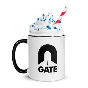 May include: A white ceramic mug with a black handle and rim, featuring a black graphic of an archway with the word "GATE" below. The mug is topped with whipped cream, blue sprinkles, and a red and white striped straw.