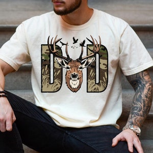 May include: A white t-shirt with a camouflage print design featuring a deer head and the word "DAD".