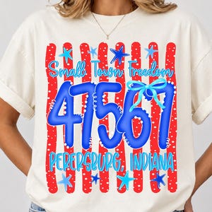 May include: Cream-coloured t-shirt with a patriotic design. The design features red and white stripes, blue stars, and the text "Small Town Freedom 47561 Petersburg, Indiana" in blue and light blue.