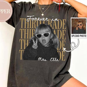 Może przedstawiać: Ciemnoszary t-shirt oversize z nadrukiem graficznym przedstawiającym dziecko w okularach przeciwsłonecznych wykonujące znak pokoju. Tekst głosi Forever in THIRD GRADE oraz Mrs. Ella. Mała ramka pokazuje oryginalne zdjęcie i polecenie przesyłania.