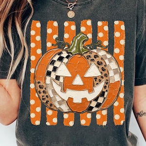 May include: A charcoal gray t-shirt featuring a central graphic of a jack-o'-lantern pumpkin. The pumpkin has a textured, painted appearance with segments of black and white checkerboard and leopard print patterns over orange vertical stripes.