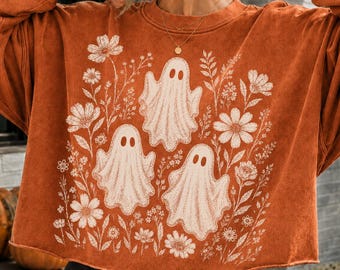 May include: A burnt orange long-sleeve sweatshirt featuring a cream-colored graphic print of three ghosts surrounded by various wildflowers and foliage. The garment has a crew neckline, long sleeves, and a raw-edge hemline at the waist.