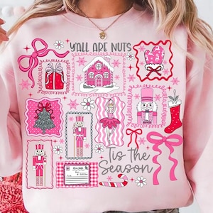 May include: A pale pink sweatshirt featuring a Christmas-themed design. The print includes pink images of a gingerbread house, nutcrackers, a Christmas tree, and the text "Y'all Are Nuts" and "Tis the Season".