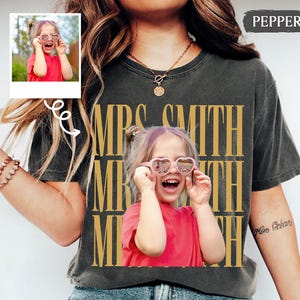 Pode incluir: Uma t-shirt cinza-carvão de manga curta com uma grande impressão fotográfica centralizada de uma criança a usar óculos de sol em forma de coração e uma t-shirt vermelha. O texto "MRS. SMITH" está impresso numa fonte dourada vertical.