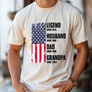 K&ouml;nnte beinhalten: Cremefarbenes T-Shirt mit amerikanischer Flagge und dem Text "LEGEND SINCE 1975, HUSBAND SINCE 1995, DAD SINCE 1998, GRANDPA SINCE 2025" in Schwarz. Das T-Shirt hat kurze &Auml;rmel und einen Rundhalsausschnitt.
