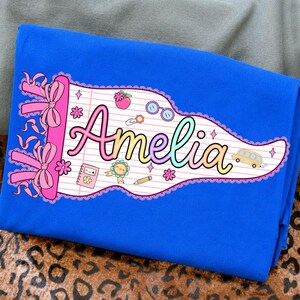 May include: A bright blue t-shirt featuring a pennant-shaped graphic with a lined paper background. The name Amelia is written in a colorful gradient font. The design includes pink bows, a school bus, a notebook, a pencil, and small flowers.