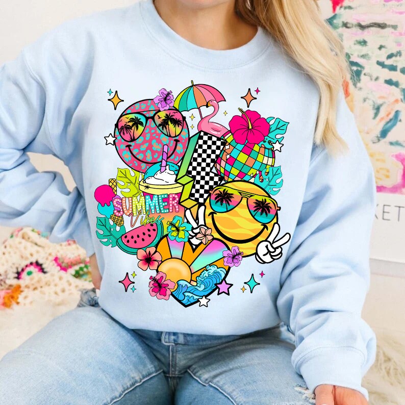 Peut inclure: Un sweat-shirt bleu clair avec un imprim&eacute; graphique color&eacute; repr&eacute;sentant deux visages souriants portant des lunettes de soleil, des palmiers, une past&egrave;que, un flamant rose, un parasol de plage et le texte "Summer Vibes".