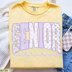 May include: Butter yellow t-shirt with the word "SENIOR" in large, decorative letters. The letters are filled with floral, striped, and bow patterns in shades of purple and white. The text "twenty twenty-seven" is written below in a cursive font.