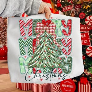 May include: White Christmas-themed tote bag featuring the words "MERRY CHRISTMAS" in a festive design. The design includes a Christmas tree, snowflakes, and a large plaid bow. The bag is held by a person.
