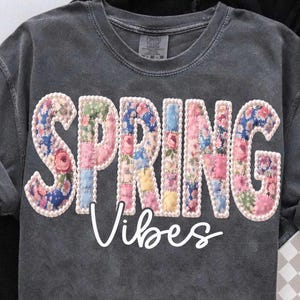 Könnte beinhalten: Dunkelgraues Comfort Colors T-Shirt mit den Worten "SPRING Vibes" in einem floralen Patchwork-Design. Die Buchstaben sind mit einer weißen Perlenborte umrandet, und das Wort "Vibes" ist in weißer Schrift geschrieben.