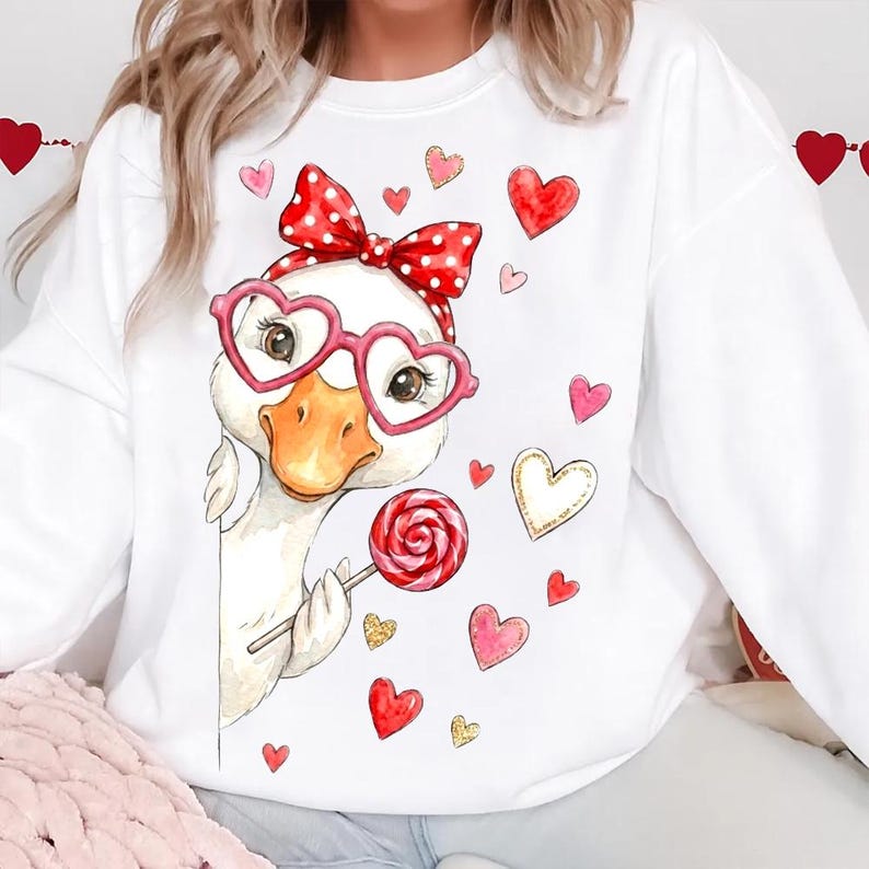 May include: White sweatshirt featuring a cartoon duck wearing heart-shaped glasses and a red polka dot bow. The duck is holding a lollipop and surrounded by red, pink, and gold hearts. A Valentine's Day themed design.