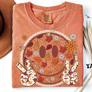 May include: A peach-colored Comfort Colors t-shirt featuring a smiley face design with fall leaves and flowers. The smiley face has peace signs with leaf patterns. The shirt has a round neckline and a small tag that says "Comfort Colors".