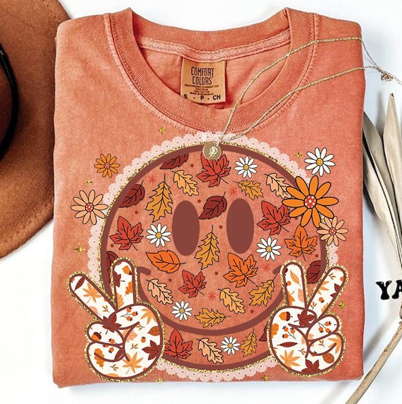 May include: A peach-colored Comfort Colors t-shirt featuring a smiley face design with fall leaves and flowers. The smiley face has peace signs with leaf patterns. The shirt has a round neckline and a small tag that says "Comfort Colors".