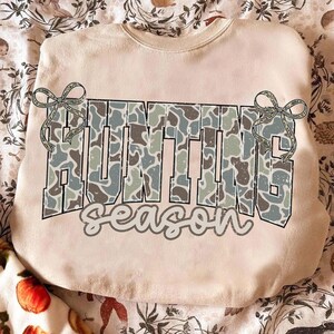May include: A beige sweatshirt with the words "HUNTING season" in a camouflage pattern. The word "HUNTING" is in large block letters, and "season" is in a script font. Two decorative bows are on either side of the word "HUNTING".