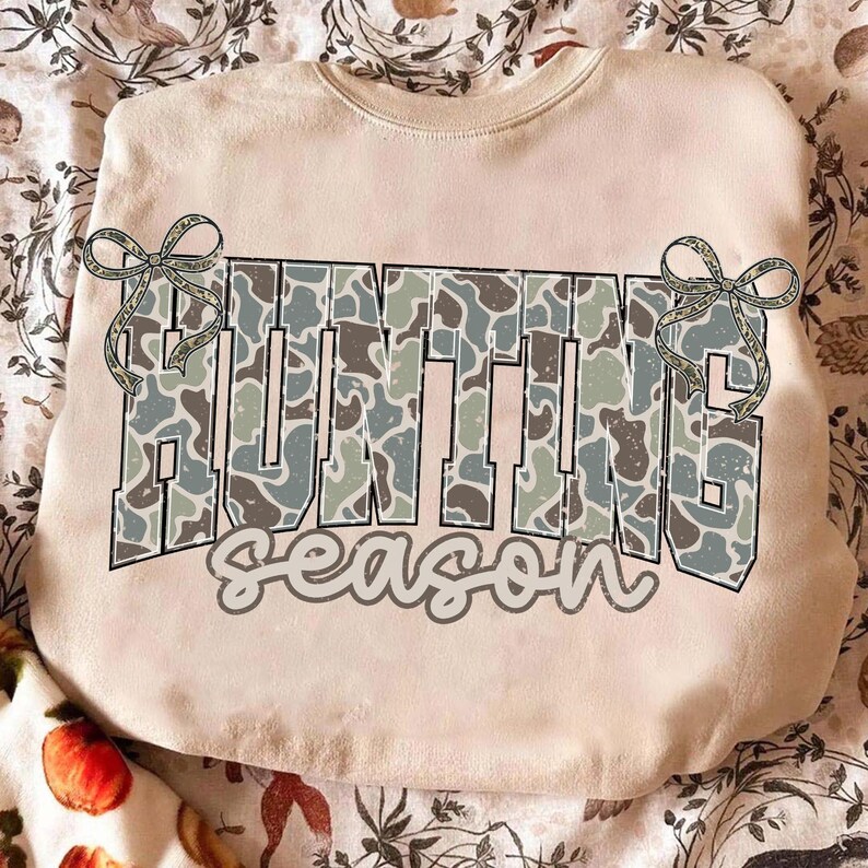 May include: A beige sweatshirt with the words "HUNTING season" in a camouflage pattern. The word "HUNTING" is in large block letters, and "season" is in a script font. Two decorative bows are on either side of the word "HUNTING".