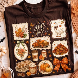 K&ouml;nnte beinhalten: Ein dunkelbraunes T-Shirt mit einer Collage aus herbstlichen Illustrationen, darunter eine Gans mit karierter Schleife, ein orangefarbener Pickup mit K&uuml;rbissen, ein Blumenstrau&szlig;, Zimtschnecken, ein Kaffeebecher und der Text Hello fall.