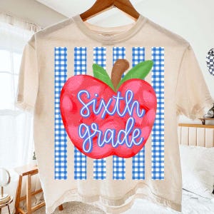 May include: A cream-colored short-sleeve t-shirt featuring a graphic of a red apple with green leaves. The apple is centered over a blue and white gingham pattern. The text "Sixth grade" is written in blue script across the center of the apple.