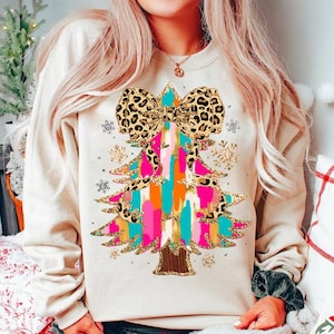 May include: A cream-colored sweatshirt with a colorful Christmas tree design. The tree is decorated with a leopard print bow, gold glitter, and colorful stripes. The tree is surrounded by snowflakes.